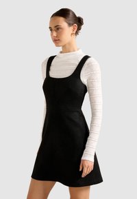 Femme aux cheveux noirs en chignon, portant des boucles d'oreilles en or, un haut blanc à manches longues texturé et une robe noire sans manches, sur un fond uni.