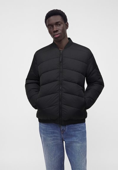 Jeune homme portant une veste matelassée noire zippée et un jean bleu, debout les mains dans les poches sur un fond clair uni.