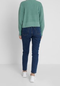 Hellgrüner gestrickter Pullover mit geripptem Muster, kombiniert mit blauen Denim-Jeans mit Gesäßtaschen und schmal zulaufendem Bein. Weiße Sneakers.