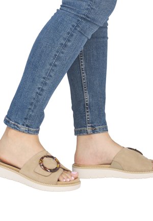 Beine in blauen Jeans, die beige Schiebe-Sandalen mit großer Schildpatt-Schnalle und weißen Sohlen tragen, Zehen in blassrosa lackiert.