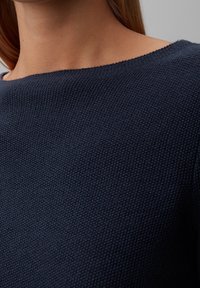 Zbliżenie osoby w ciemnoniebieskim, teksturowanym swetrze z szerokim dekoltem, ukazującym szyję i okolice ramion.