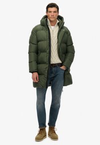 Hombre con abrigo acolchado largo verde, suéter de punto crema, jeans azules, cinturón marrón, botas beige, reloj de pulsera, de pie con una mano en el bolsillo.