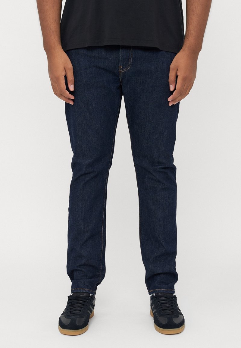 Homme portant un jean bleu foncé, des baskets noires avec semelles en caoutchouc, et un t-shirt noir, debout devant un fond clair uni.