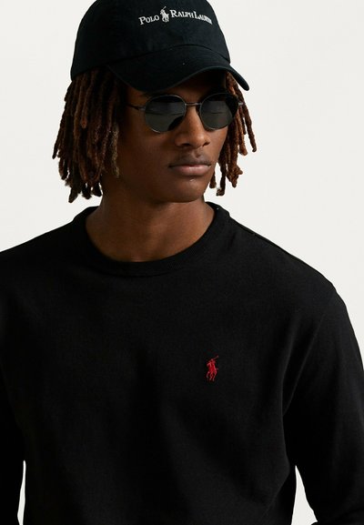 La maglietta nera a maniche lunghe presenta un piccolo logo ricamato rosso. Abbinata a un cappellino nero e occhiali da sole neri rotondi, mostra un look informale.