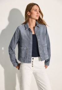 Chaqueta a rayas azul y blanca con diseño de cuello, dos bolsillos en la parte frontal y cierres de presión metálicos, combinada con una camiseta sin mangas azul marino y pantalones blancos.