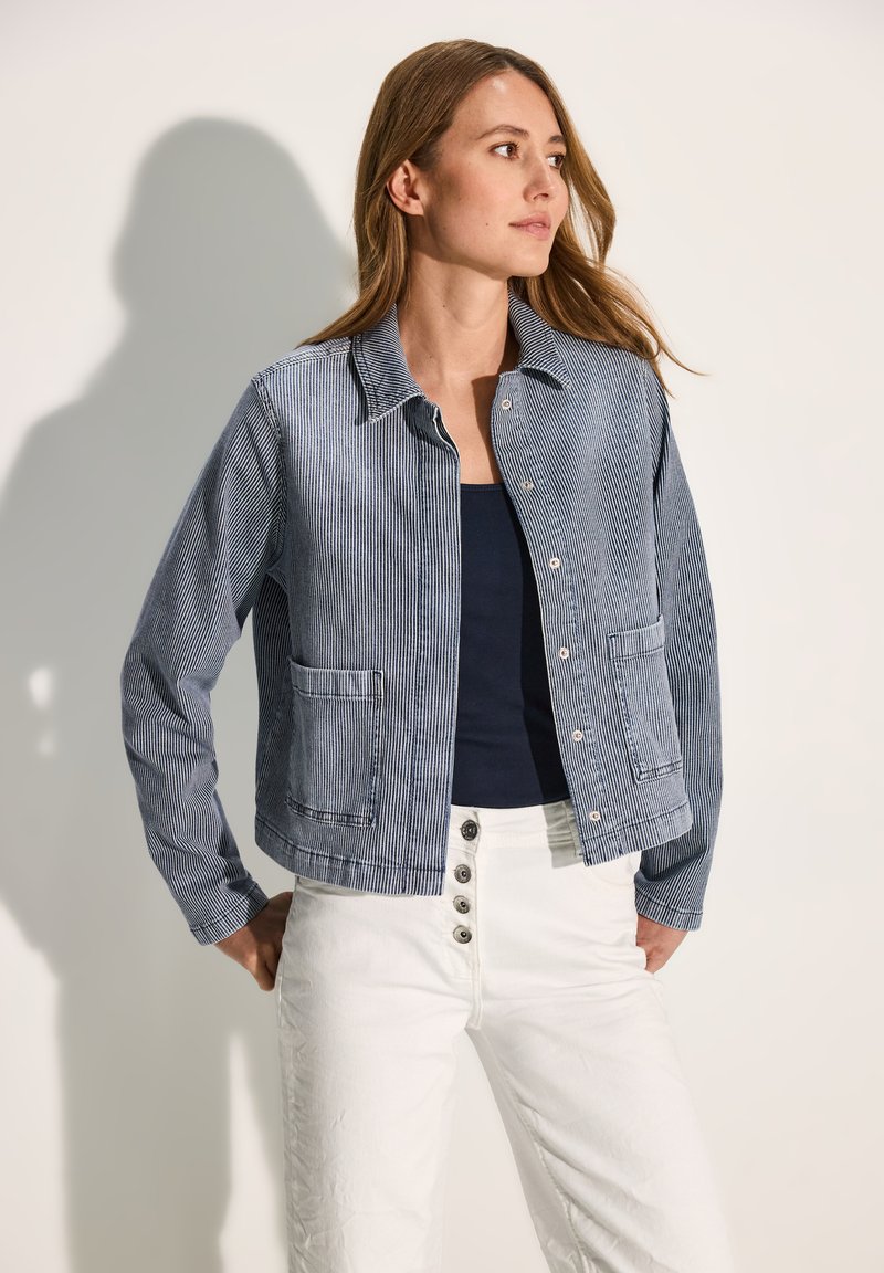 Chaqueta a rayas azul y blanca con diseño de cuello, dos bolsillos en la parte frontal y cierres de presión metálicos, combinada con una camiseta sin mangas azul marino y pantalones blancos.