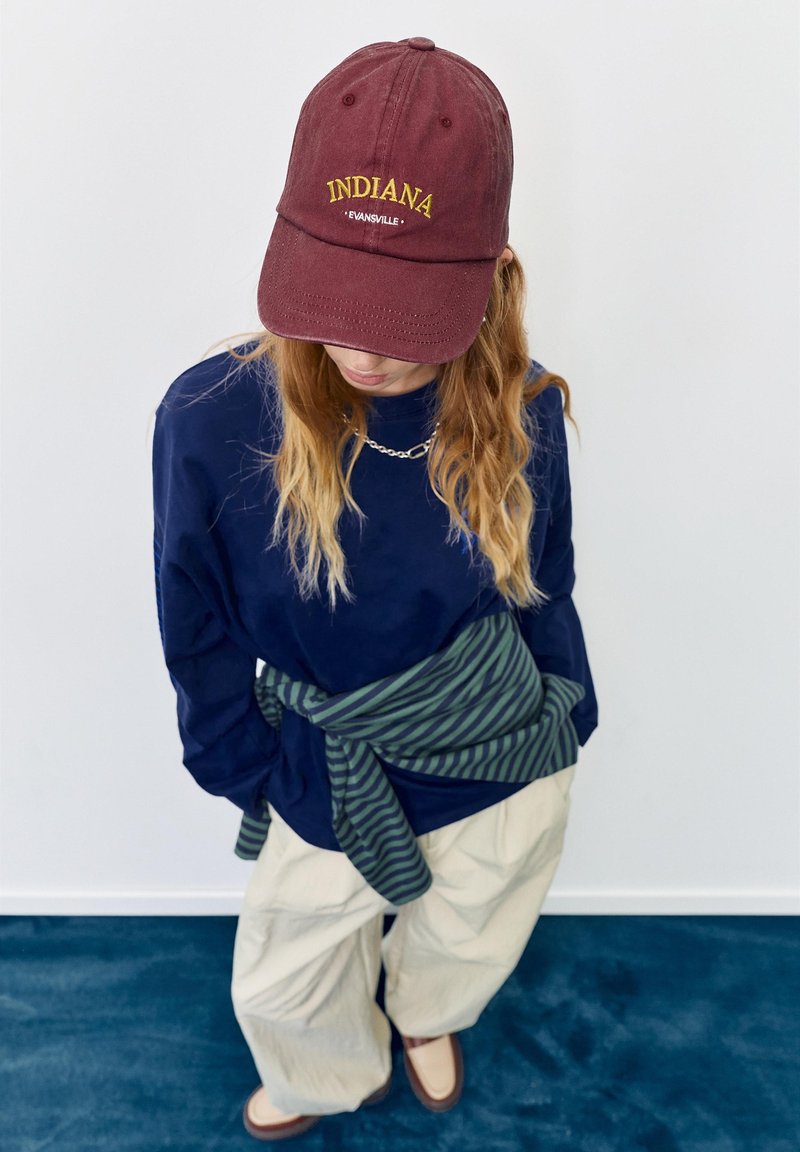 Personne regardant vers le bas portant une casquette de baseball Indiana bordeaux, une chemise bleu marine, un pantalon beige, un pull vert rayé noué autour de la taille, et un collier.