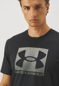 Svart T-skjorte laget av bomull, med et stort grått Under Armour-logo på framsiden. Korte ermer og rund hals.