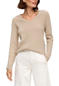 Pull beige en tricot avec col en V, manches longues, texture côtelée et fentes sur les côtés, associé à un pantalon blanc taille haute.