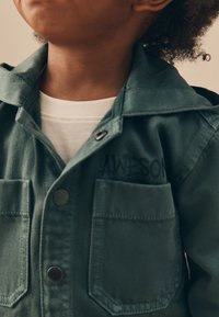 Jachetă verde din denim cu guler pliabil, două buzunare frontale, nasturi metalici și cuvântul „AWESOM” imprimat pe piept. Cămașă albă dedesubt.