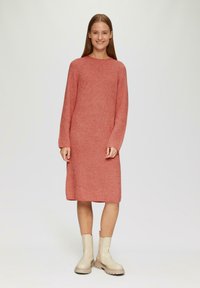 Strickkleid in sanftem Koralle, mit lockerer Passform, langen Ärmeln und geradem Saum. Kombiniert mit cremefarbenen Stiefeletten.
