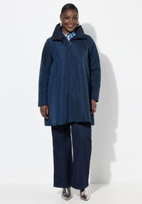 Cappotto impermeabile blu navy con colletto alto, cerniera frontale e tasche laterali, indossato sopra pantaloni larghi scuri e stivali alla caviglia metallici.