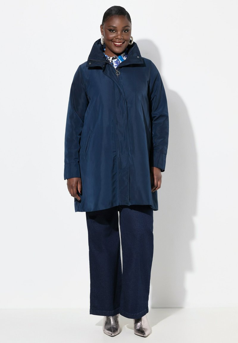 Cappotto impermeabile blu navy con colletto alto, cerniera frontale e tasche laterali, indossato sopra pantaloni larghi scuri e stivali alla caviglia metallici.