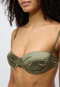 Σουτιέν από σατέν σε ελιά χρώμα με ντεκολτέ καρδιάς, ενίσχυση με μπανέλα, ρυτιδωτές λεπτομέρειες και ρυθμιζόμενους ιμάντες. Διαθέτει μικρή διακριτική λογότυπο.