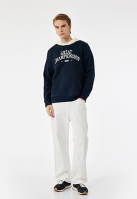 Sudadera con capucha en color navy con impresión "GRAN CAMPEONATO", bolsillo frontal, combinada con pantalones anchos blancos y zapatillas navy. Textura suave.