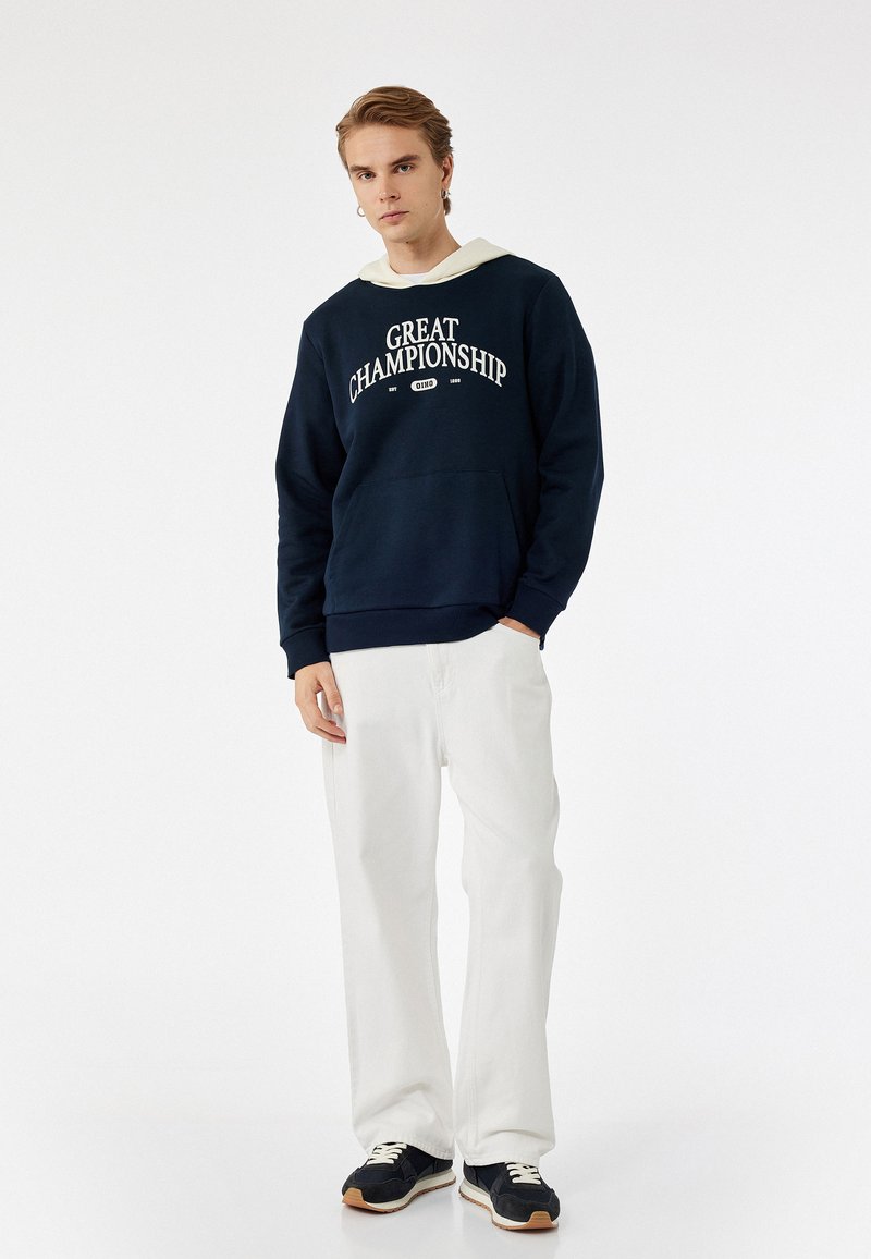 Sudadera con capucha en color navy con impresión "GRAN CAMPEONATO", bolsillo frontal, combinada con pantalones anchos blancos y zapatillas navy. Textura suave.