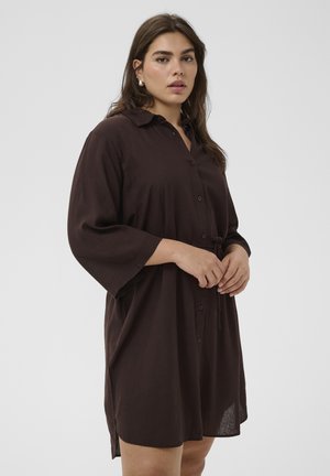 Frau mit langem dunklem Haar, die ein lockeres, dunkelbraunes Hemdblusenkleid mit dreiviertel Ärmeln trägt und die Hände nach vorn hält.