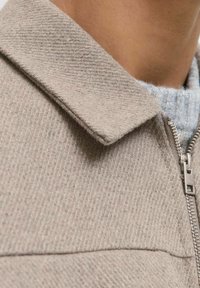 Svijetlosiva jakna na zip s teksturiranim tkanim uzorkom, oštrim ovratnikom i metalnim patentnim zatvaračem. Blizu prikaz mekog materijala.