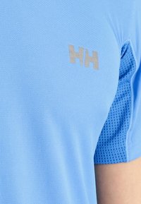 Ljusblå atletisk tröja med strukturerad tyg, nätpaneler och en silverfärgad logotyp på vänster bröstområde. Korta ärmar med platta sömmar.