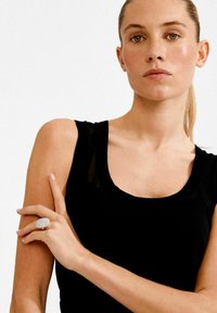 Hõbesõrmus tekstuurse ovaalse pinnaga, kantud sõrmel. Modell kannab musta varrukateta topi ja neutraalset küünelakki, taustaks heledat värvi pind.