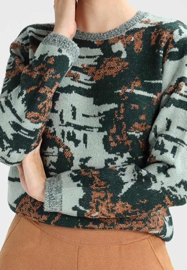Personne portant un pull ras-du-cou avec des motifs métalliques abstraits verts, gris et cuivre, associé à un pantalon marron taille haute.