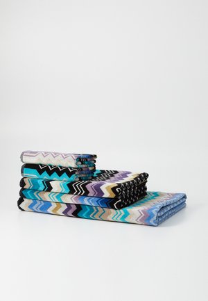 Serviettes colorées à motifs empilées de différentes tailles. Présentent des motifs en zigzag dans des teintes de bleu, violet, noir et beige sur un fond clair.