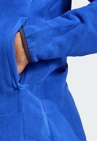 Blue fleece jacket featuring a side zip pocket and contrasting dark cuff trim. Surface shows a soft, textured finish.
