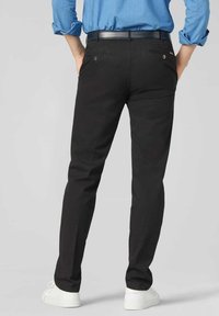 Pantalons noirs en tissu lisse, avec une coupe ajustée, des passants de ceinture et deux poches arrière avec des boutons décoratifs. Portés avec des chaussures blanches.