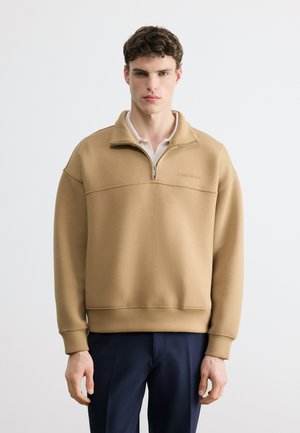 Junger Mann trägt einen beigen Sweatshirt mit Viertel-Reißverschluss über einem weißen Hemd und dunkelblauen Hosen, steht vor einem schlichten hellen Hintergrund.