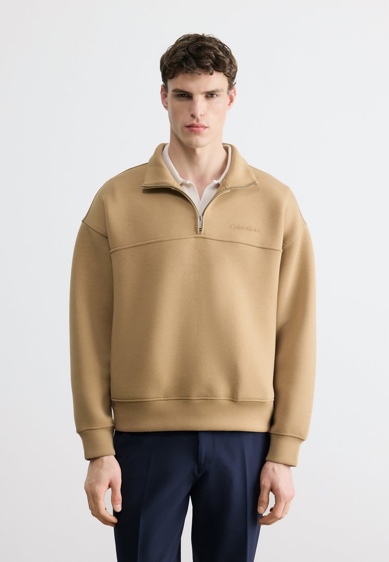 Νεαρός άνδρας που φοράει μπεζ φούτερ με φερμουάρ ως το στήθος πάνω από λευκό πουκάμισο και σκούρο μπλε παντελόνι, στέκεται μπροστά σε έναν απλό ανοιχτόχρωμο φόντο.