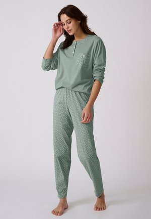 Femme debout pieds nus portant un haut vert sauge à manches longues et un pantalon de pyjama à pois, main touchant les cheveux, regard vers le bas.