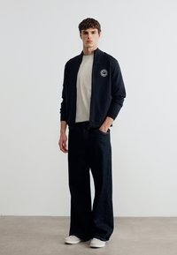 Νέος άνδρας όρθιος, φορώντας μαύρο μπουφάν με φερμουάρ και εμβληματικό λογότυπο, μπεζ πουκάμισο, φαρδιά μαύρη παντελόνια και λευκά αθλητικά παπούτσια, σε απλό λευκό φόντο.