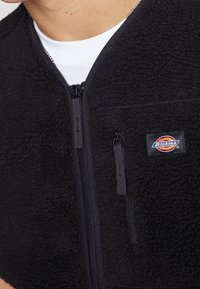 Gilet in pile nero con chiusura a zip, dotato di un logo in tessuto e una superficie liscia e testurizzata. Include una piccola tasca laterale con zip.