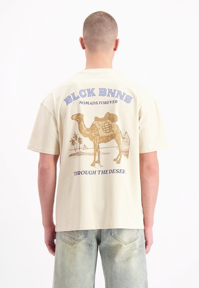 Crème katoenen T-shirt met een gouden kameelafbeelding, palmbomen en piramides, met de tekst "BLCK BNNS" en "THROUGH THE DESERT" in het blauw.