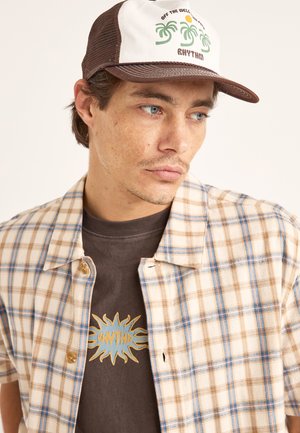 Camisa de manga corta a cuadros beige y azul sobre una camiseta marrón con un gráfico de sol, combinada con una gorra de béisbol marrón de malla.