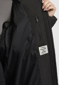 Eine Hand hält eine schwarze Jacke offen und zeigt die Innentasche mit Reißverschluss und einem Stoffetikett, auf dem steht: "Eine warme Umarmung aus Stoff."