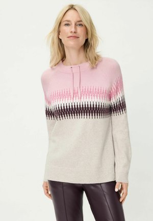 Hellrosa Pullover mit einem Farbverlauf von dunkelrosa zu schwarzen Zickzackmustern. Weiche Textur, Rundhalsausschnitt und gerippte Bündchen. Kombiniert mit dunkler Hose.
