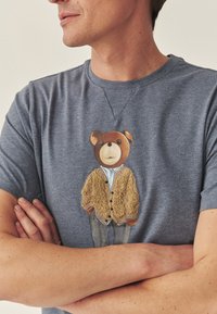 Šedo-modré bavlněné tričko s motivem medvěda v žlutém svetříku přes bílou košili a šedé kalhoty. Krátké rukávy a kulatý výstřih.