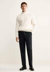 Maglione beige chiaro a coste con collo alto abbinato a pantaloni scuri a righe sottili e scarpe nere. L'outfit presenta una vestibilità comoda e una texture liscia.