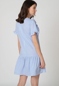 Mujer con un vestido corto de cuadros en azul claro y blanco, con mangas y dobladillo con volantes, de pie de lado contra un fondo liso.
