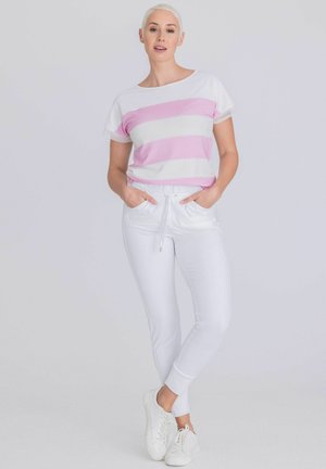 Femme debout les mains dans les poches portant un haut rayé rose et blanc à manches courtes, un pantalon blanc et des baskets blanches sur fond gris.