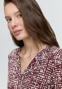 Detailní záběr ženy s dlouhými, vlnitými hnědými vlasy, oblečené v vzorované vínové blůze, která se mírně dívá na stranu na neutrálním pozadí.