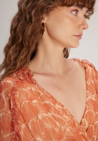 Orangefarbene Bluse mit Rüschendetail am Kragen. Weicher, leichter Stoff mit goldenen Akzenten, der Textur und Design hervorhebt.