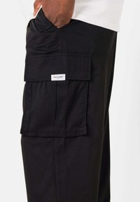Zwarte cargobroek met een voorkantzak met klep, zichtbare stiksels en een merklabel. Gemaakt van stevig materiaal met een ontspannen pasvorm.