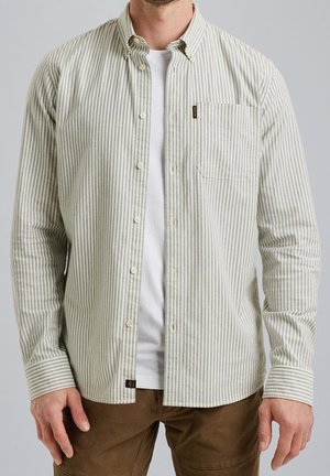 Homme portant une chemise beige et blanche à rayures verticales boutonnée sur un t-shirt blanc avec un pantalon marron, debout devant un fond uni.
