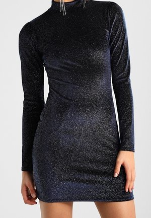 Eng anliegendes, langärmeliges, schwarzes glitzerndes Minikleid, getragen von einer stehenden Person mit baumelnden Kristallohrringen.