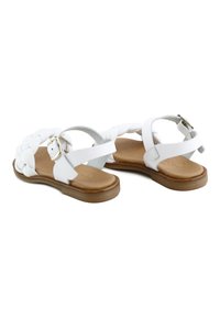 Sandalias de cuero blanco con tira trenzada en el frente, hebilla en el tobillo y suela plana marrón, mostradas desde ángulos trasero y lateral.