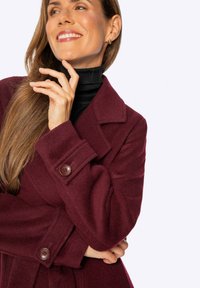 Abrigo de lana burdeos con un cuello estructurado, que presenta dos grandes botones en el frente y una tela texturizada. El cuello alto debajo es negro.