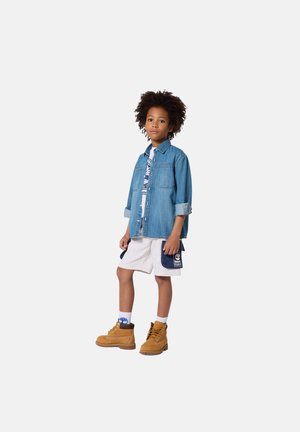 Chemise en denim, t-shirt blanc à motif graphique, short gris avec poches, et bottes beige. Cheveux courts et bouclés, debout devant un fond uni.
