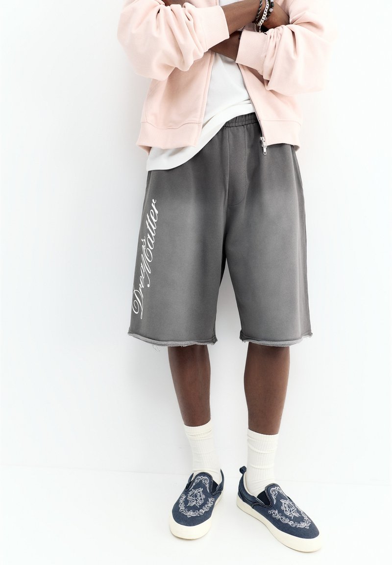 Personne portant un short gris avec le texte « Dream Matter », des chaussettes blanches, des chaussures bateau bleu marine, une veste rose clair et une chemise blanche, debout, les bras croisés.